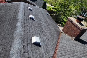 1000039159 - Integrity 1st Roofing View of a gray shingle pitched roof from above with multiple white square rooftop vents and a brick chimney on the right, plus green trees in the yard beyond.