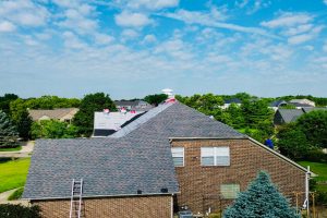 Fan 2 - Integrity 1st Roofing Brick house undergoing roof work; a ladder rests against the roof and a worker is on the far side under a sunny blue sky.