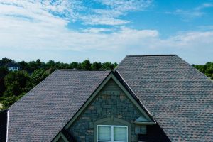 Fan6 - Integrity 1st Roofing Aerial view of gray shingle roofs and a stone-clad house under a bright blue sky.