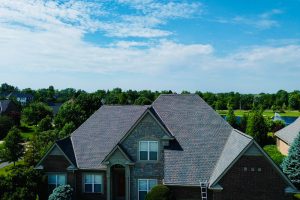 Fan7 - Integrity 1st Roofing Two-story brick-and-stone suburban house with a steep gray roof, surrounded by green trees under a blue sky.