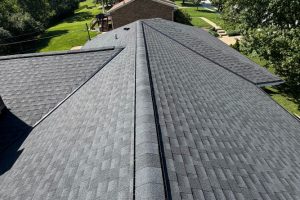 item_0ed6a294-f407-4319-a647-6d5e5b1136c7 - Integrity 1st Roofing Aerial view of a gray asphalt-shingle roof with intersecting ridges and visible vents, surrounded by a green lawn and houses in the distance.