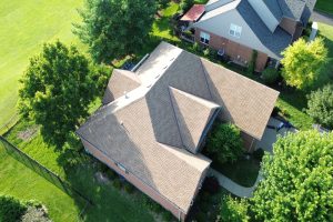 item_12afab68-1222-4abc-ace3-c812b83bfb28 - Integrity 1st Roofing Aerial view of a brick house with a tan shingle roof, surrounded by green lawn and trees, with a curved concrete walkway nearby.