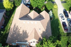 item_16341763-3d31-4e49-9ea2-d0ec4502eacc - Integrity 1st Roofing Aerial view of a large brick house with a multi-hip brown shingle roof, surrounded by green lawn and trees, with several cars parked along a curved driveway to the right.