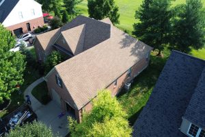 item_17df9798-81a6-4ac4-8222-2180b84f7814 - Integrity 1st Roofing Aerial view of a brick house with a tan shingled roof, neighboring homes, a curved concrete driveway, and green lawns with trees surrounding the properties.