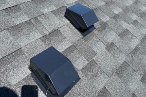 item_3789d630-1671-492f-8daa-47a4e2d800ab - Integrity 1st Roofing Two dark rooftop vent caps protruding from a gray shingled roof, viewed from above.