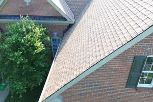 item_3dae0578-0d28-4d4e-ad99-076c334ae0cd - Integrity 1st Roofing A brick house viewed from above showing a steep tan shingle roof, a large leafy tree on the left, and a window with green shutters on the right.