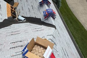 item_4fd7bd2c-f0aa-482f-b3df-c6e59076848f - Integrity 1st Roofing Rooftop underlayment visible as shingles are staged; bundles near the edge and an open cardboard box on the deck.