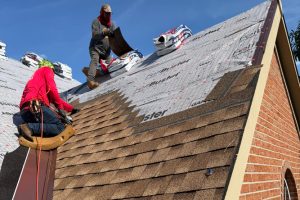 item_500e6cae-4c9e-4ca9-99fb-6e04522486c8 - Integrity 1st Roofing Two workers install brown asphalt shingles on a steep residential roof under a clear blue sky, with bundles of shingles nearby