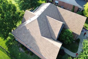item_7c0d2d91-70ab-432e-8253-6b00b8f7baba - Integrity 1st Roofing Aerial view of a large suburban house with tan shingle roof and multiple gables, surrounded by green trees and a curved sidewalk.