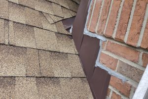 item_7d3ad7d8-a09b-4439-856f-7a1a56ec1f01 - Integrity 1st Roofing Close-up of a brown asphalt shingle roof edge next to a brick wall with metal flashing visible at the joint.