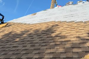 item_87f3c6aa-1c7f-4d2b-9248-f3be7fdf3ac1 - Integrity 1st Roofing Angle-view roof under construction: brown asphalt shingles on the lower slope and white underlayment/house wrap with tools visible on the upper slope against a bright blue sky