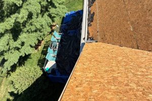 item_8f24ee15-446c-4013-a218-ded4af868835 - Integrity 1st Roofing Aerial view of a house under construction: orange plywood roof, blue tarps and portable toilets lined along the side, with trees on the left.