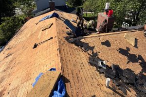 item_9145da14-53a1-4493-aa56-3674fc9315d5 - Integrity 1st Roofing Roofers repair a pitched roof with exposed plywood and torn shingles, tools scattered near a brick chimney.