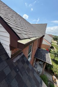 06bb7efc-2ab6-4d21-9c4c-c62884fdcf1b-item_d003a52f-d3c7-4427-a410-9e72020e1864 - Integrity 1st Roofing Aerial side view of a brick house with a dark shingle roof and brown gutter, showing a missing section near the downspout; neighboring homes and a green lawn are in view.