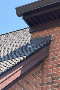3d39a408-90c6-415c-8571-72ba4d58880d-item_aefc4ef8-dd25-43ae-9e2d-2c32e451ddf8 - Integrity 1st Roofing Close-up of a brick house corner showing brown gutter and trim along a sloped asphalt shingle roof against a blue sky.