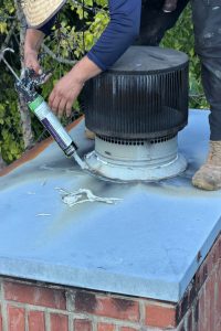 98870fbe-b138-4693-b9ca-0f6a98e90fe7-item_f6c77455-6de6-4b28-9d6f-93dde551f3a8 - Integrity 1st Roofing Worker applies sealant with a caulk gun around a roof vent on a blue parapet surface.