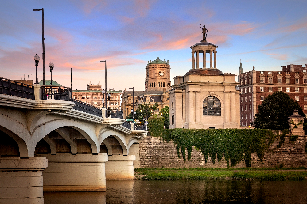 Hamilton OH - Integrity 1st Roofing Arched bridge over a river at sunset with a classical monument and clock tower in the cityscape.