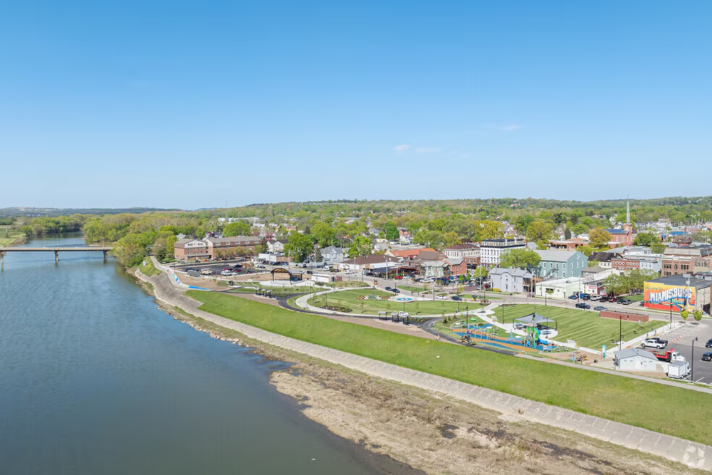 Miamisburg Ohio - Integrity 1st Roofing A riverside park with a green lawn, playground, and pathways overlooking a small town along the water; a bridge extends across the river in the distance.