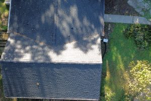 774028e7-a94b-47ee-aa5d-88cd98b5d25d-item_2983b28e-4dc3-4bc4-8459-41f41a98d39c - Integrity 1st Roofing Aerial view of a building roof with dark shingles, a lighter sloped section, and a grassy yard with a stone path along the right side.