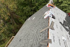 7bf8b48d-3bfd-4041-9f87-f39d9dca1c6a-item_616cc547-22e4-493d-b876-466296568ed8 - Integrity 1st Roofing A pitched roof under construction with partial dark shingles and exposed underlayment, surrounded by trees as workers pace along the ridge.