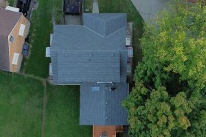 Roof Replacement014 - Integrity 1st Roofing Aerial view of a large, gray-roofed house with multiple sections, flanked by a green lawn to the left and dense trees to the right.