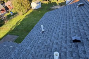 Roof Replacement05 - Integrity 1st Roofing Aerial view of a gray shingled roof with vent pipes and a small vent, with a green backyard and shed visible in the background to the left.