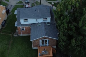 Roof Replacement06 - Integrity 1st Roofing Aerial view of a brick house with a gray roof, back wooden deck, and surrounding green lawn and trees to the right.