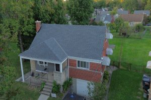 Roof Replacement11 - Integrity 1st Roofing Aerial view of a brick house with a blue-gray roof, front porch, and concrete steps, surrounded by green lawn and trees.