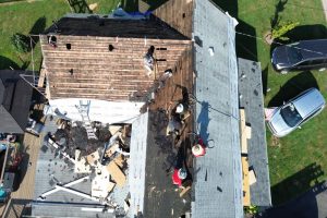 fb2ea6fd-cc09-4a1c-8772-9e6fd1f80030-item_38645b1c-73ab-4b1f-a921-488b403dbb05 - Integrity 1st Roofing Aerial view of workers on a pitched roof repairing shingles, with debris, ladders, and vehicles in the yard nearby.